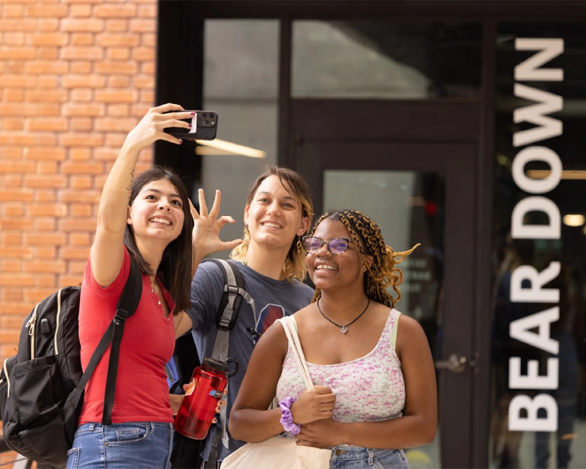 Tres estudiantes tomándose una selfie afuera del edificio del campus de la UA